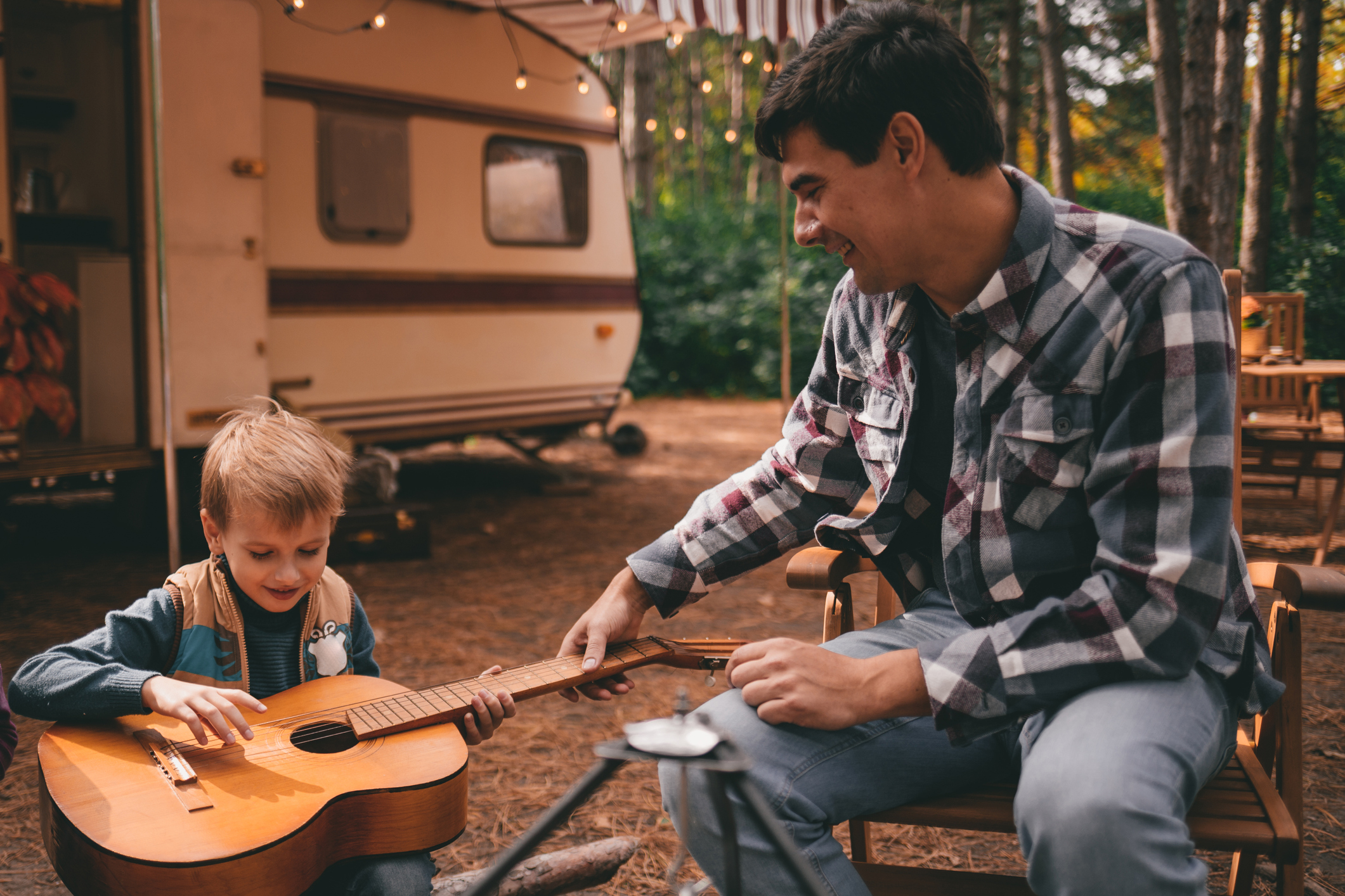 Father teaches son play guitar on camping trip relaxing in the autumn forest.