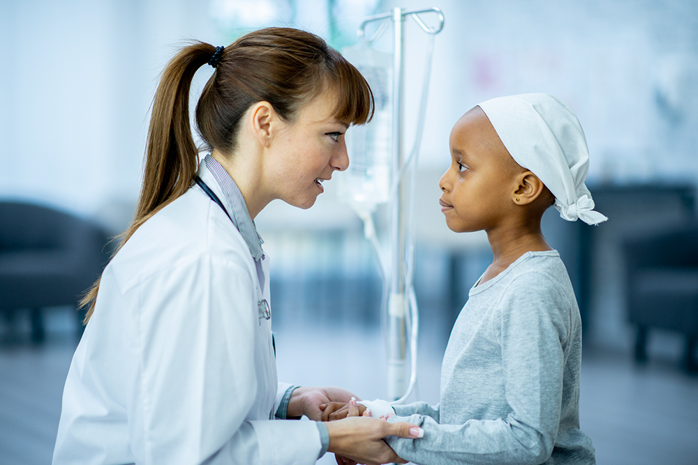 A female doctor and cancer patient are indoors in a hospital room. The doctor is holding the girl's hands to comfort her.